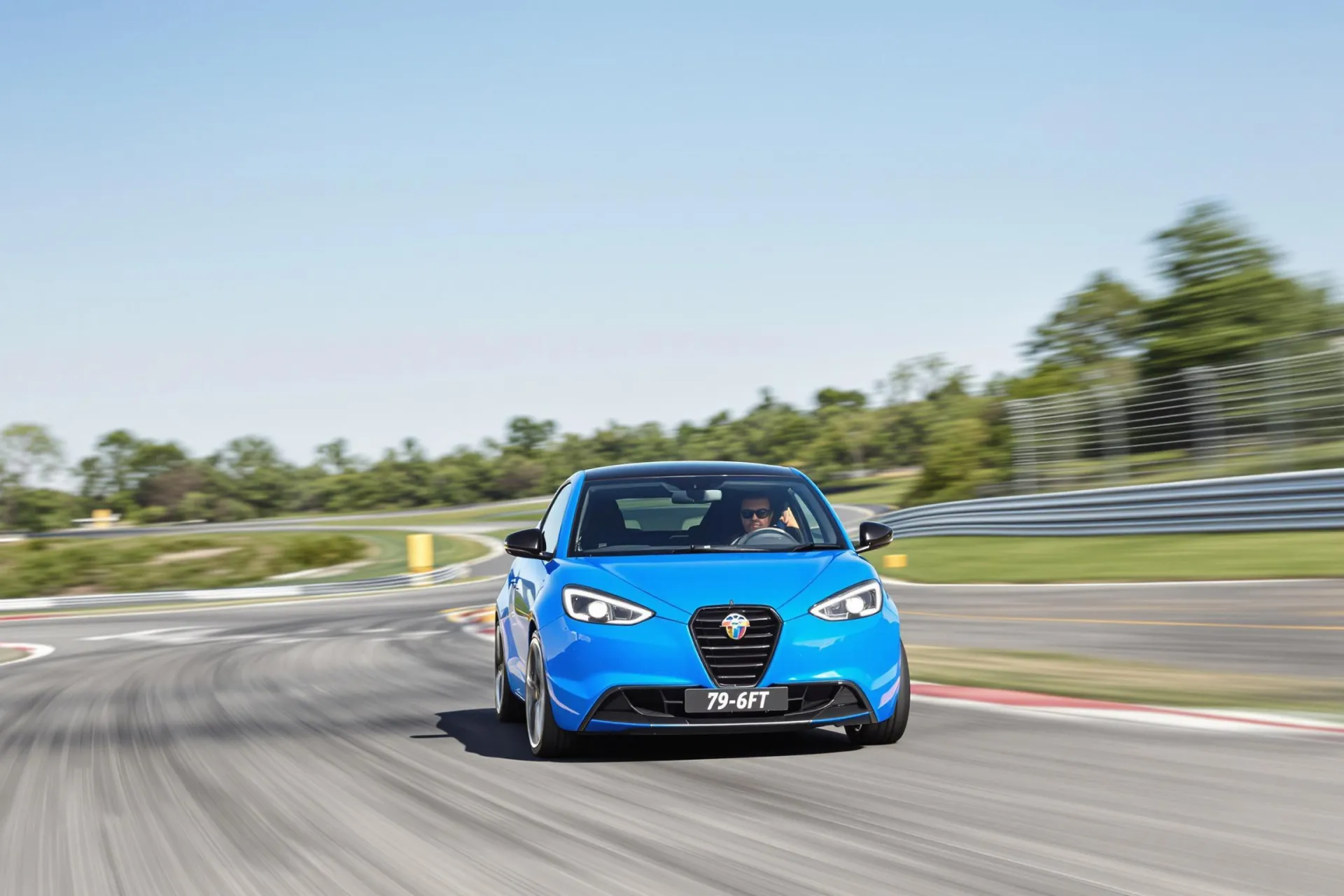 Une Lancia Ypsilon HF électrique bleue prend un virage sur circuit sous un ciel clair.