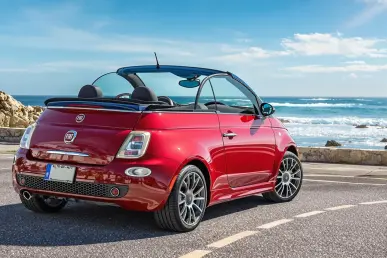 Une Fiat 500e Cabrio rouge avec toit ouvert sur une route côtière pittoresque.