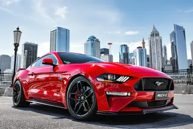 Une Ford Mustang Mach-E rouge garée devant une skyline urbaine sous un ciel bleu.
