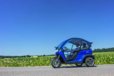 Un tricycle électrique Line bleu vibrant roulant sur une route de campagne française, entouré de champs verts et ciel bleu.