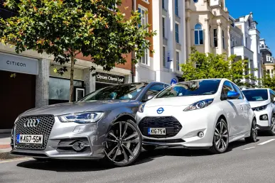 Une rue urbaine avec une Audi, une Citroën, et une Nissan Leaf sous un ciel bleu.